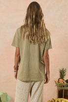 Person wearing a green shirt and white pants standing against a pink wall with a basket of fruit on a table.