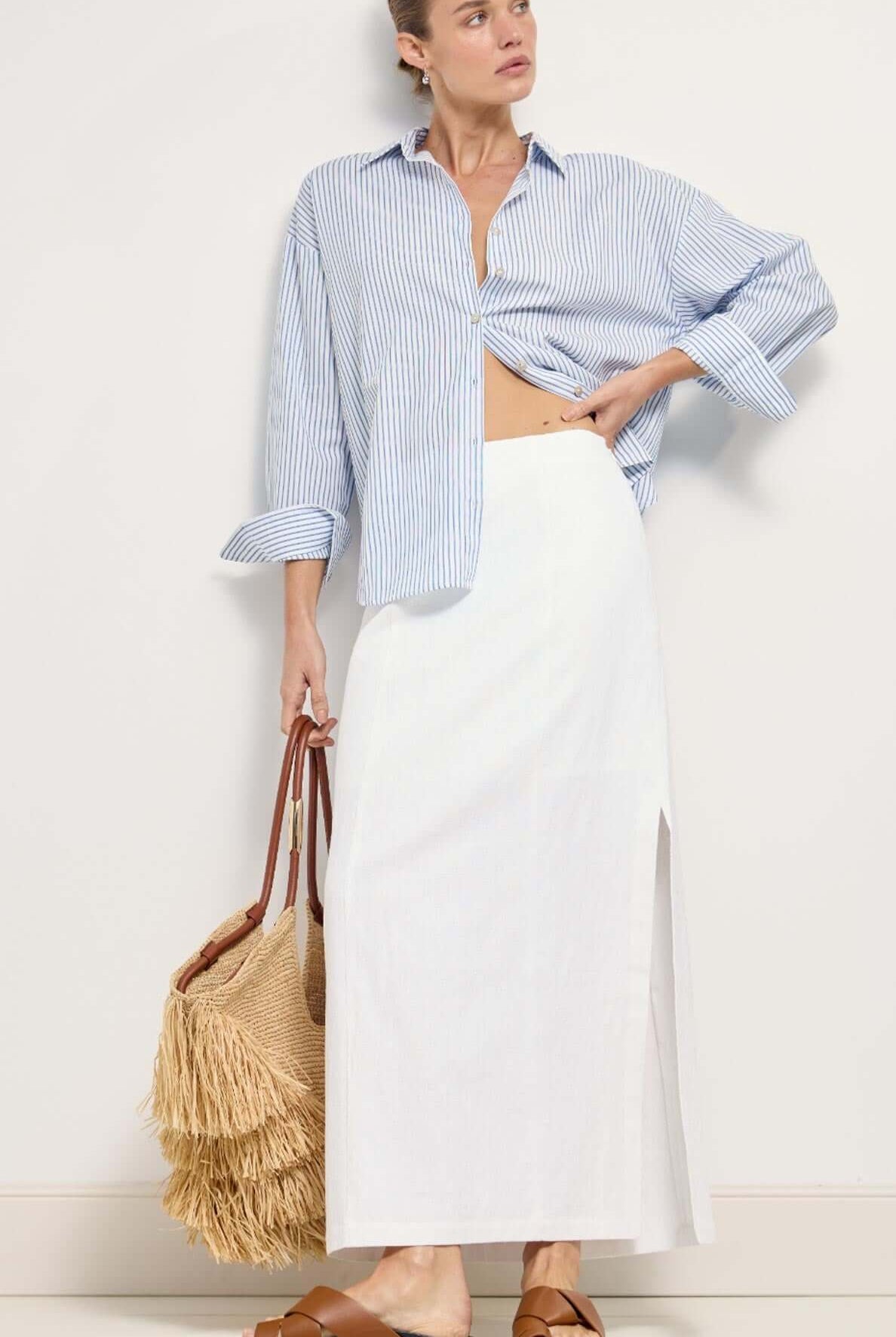 Woman wearing a blue striped shirt and white skirt, holding a woven bag against a white background