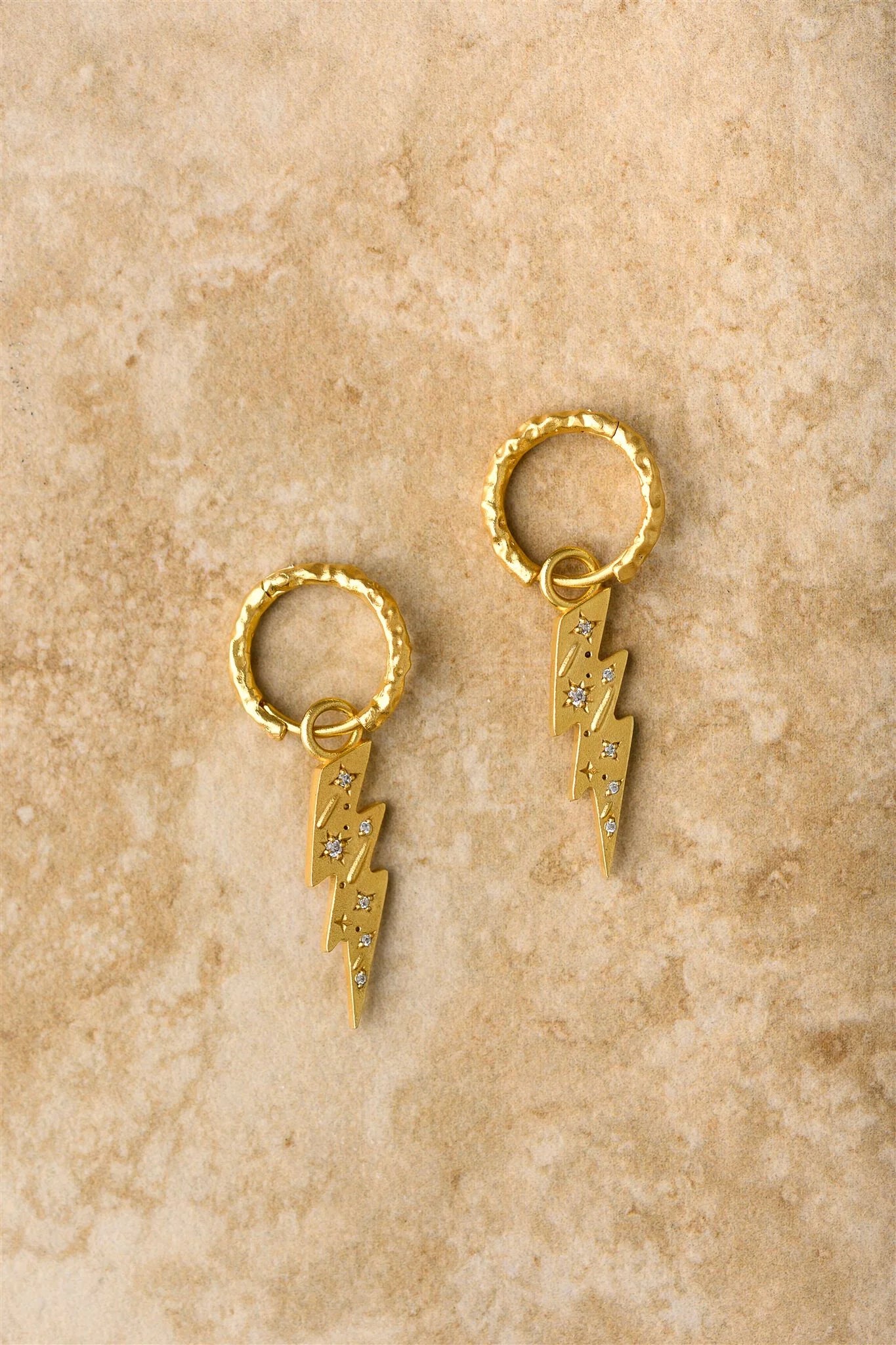 Gold hoop earrings with lightning bolt charms on a beige stone surface