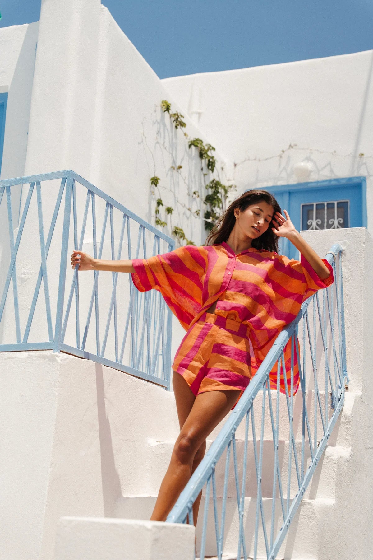 Woman in a colorful dress standing on a white staircase with blue railings against a white wall and blue sky.