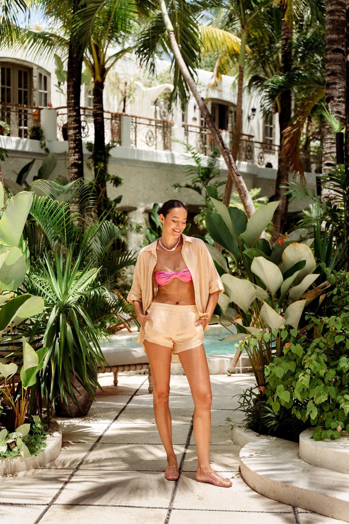 Woman in a pink bikini top and beige shorts standing in a tropical setting with palm trees and plants.