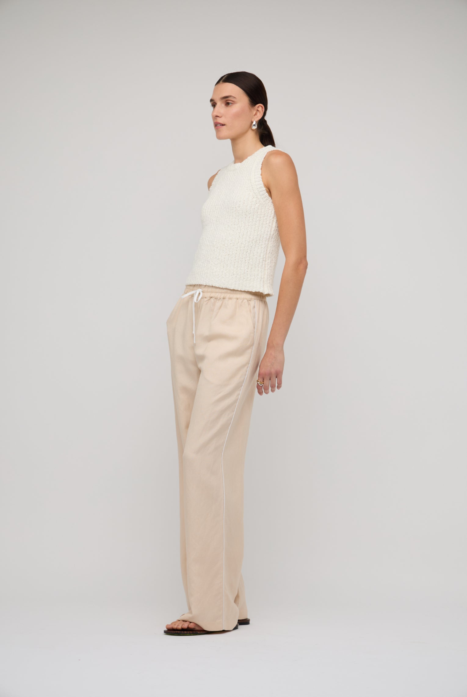 Woman wearing a white sleeveless top and beige pants on a plain background