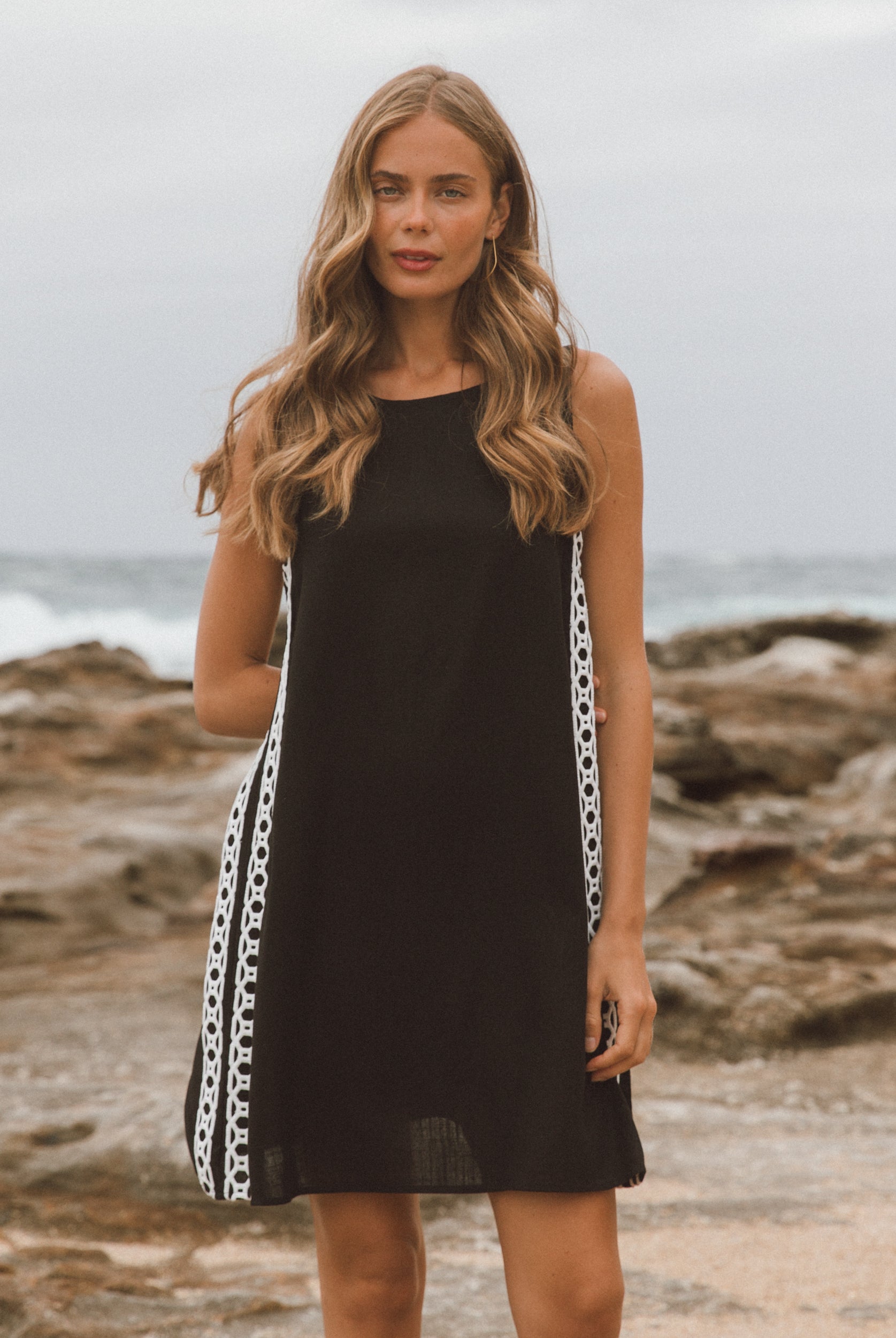Woman in a black dress with chain detail standing on a rocky beach.