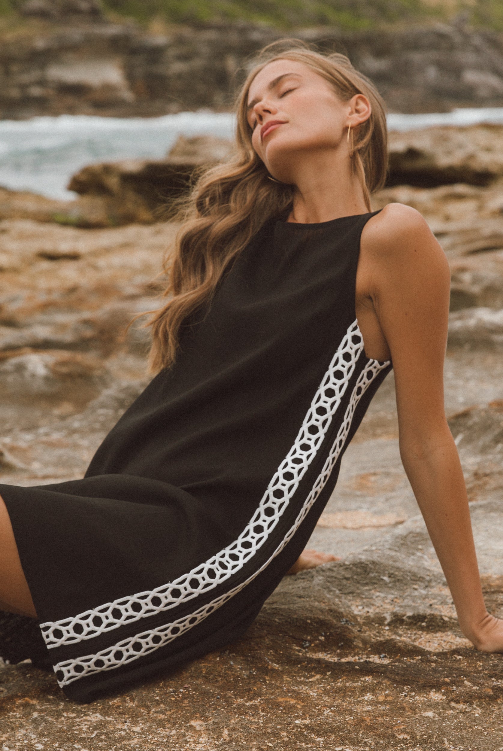 Woman in a black dress with white trim sitting on rocks by the water.