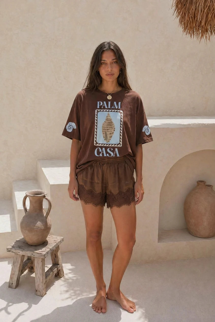 Woman wearing a brown 'Palm Casa' t-shirt and shorts in a sunlit outdoor setting.