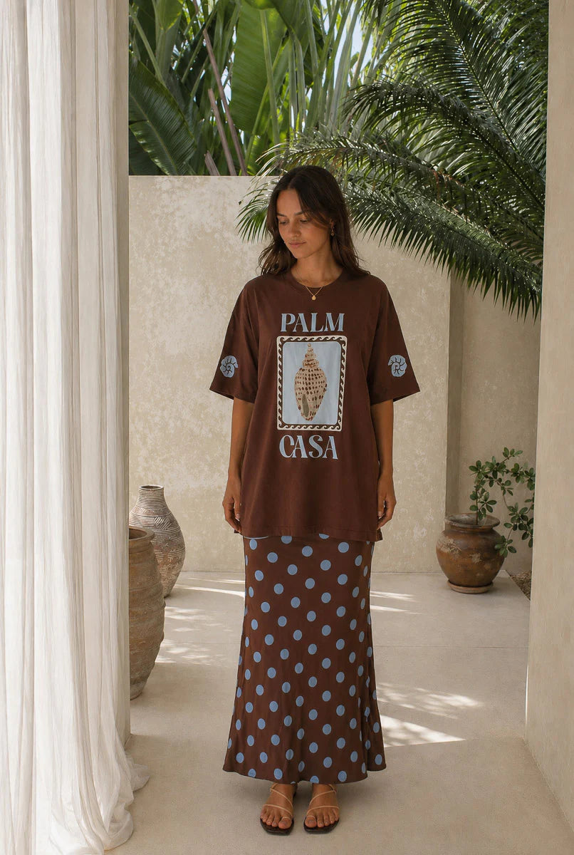 Woman wearing a brown t-shirt with 'Palm Casa' design and a patterned skirt in a tropical setting.