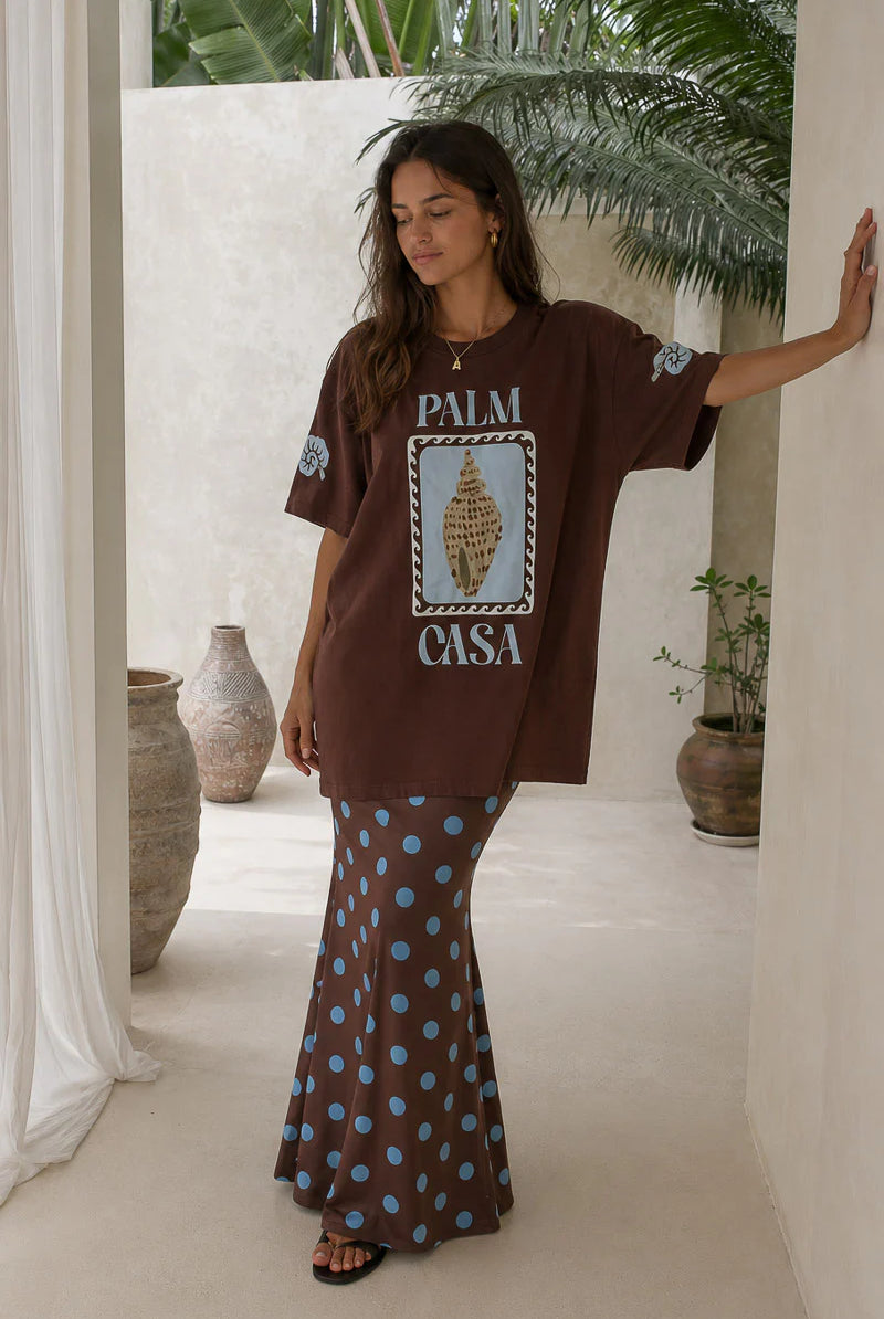 Woman wearing a brown oversized t-shirt with 'Palm Casa' design and patterned skirt in an outdoor setting.