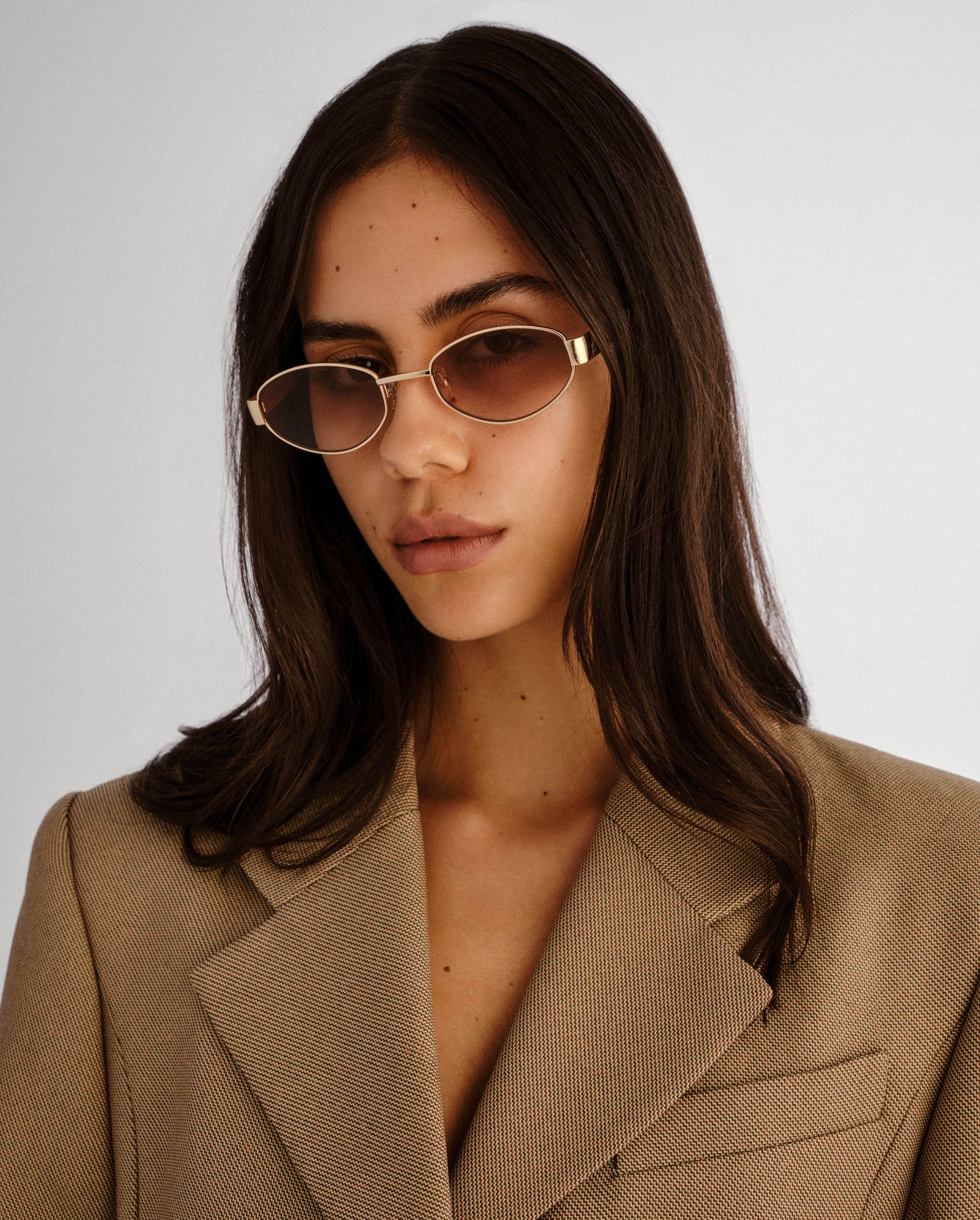 Woman wearing sunglasses and a beige blazer against a plain background