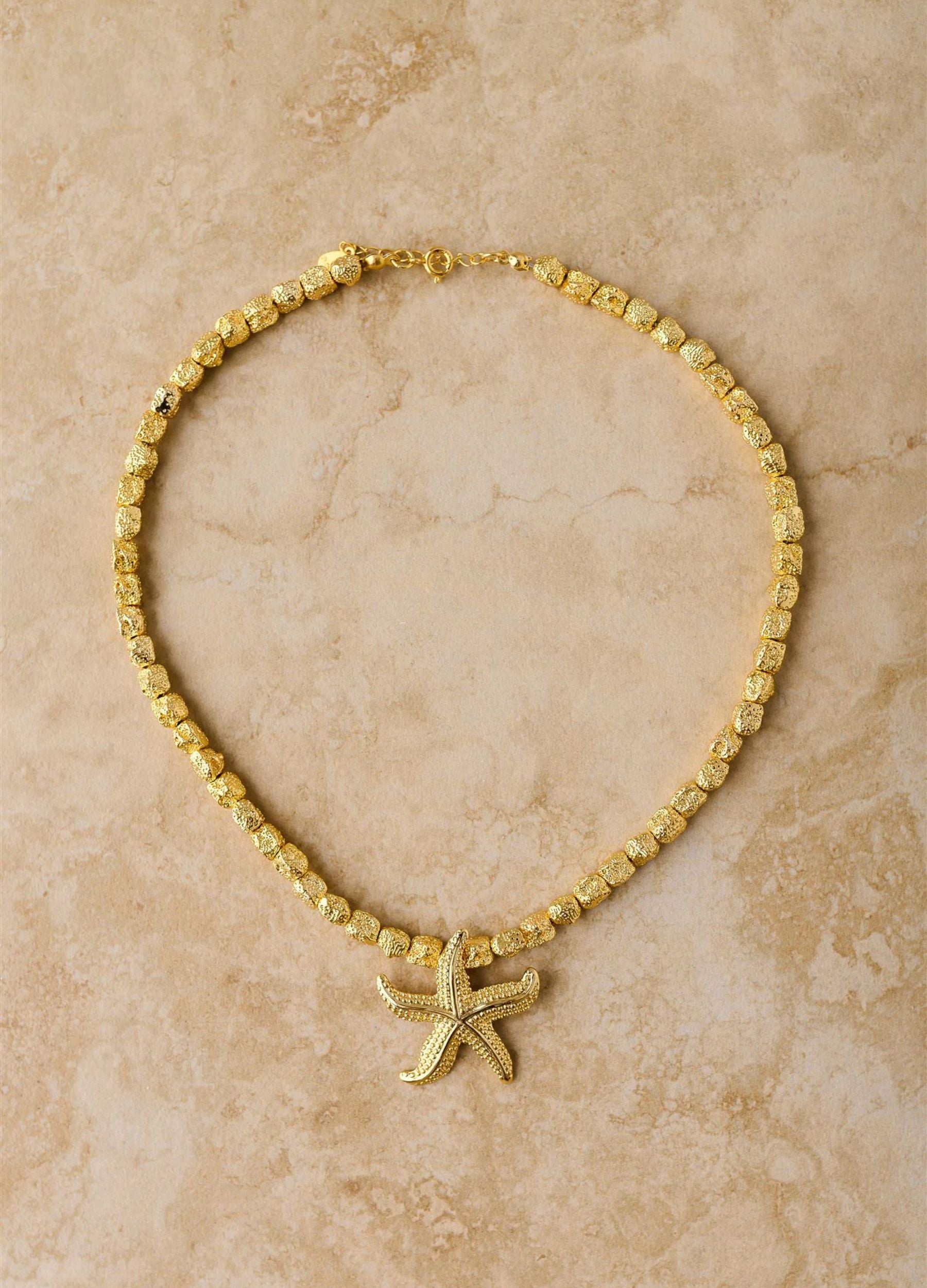 Gold beaded necklace with a starfish pendant on a beige marble background