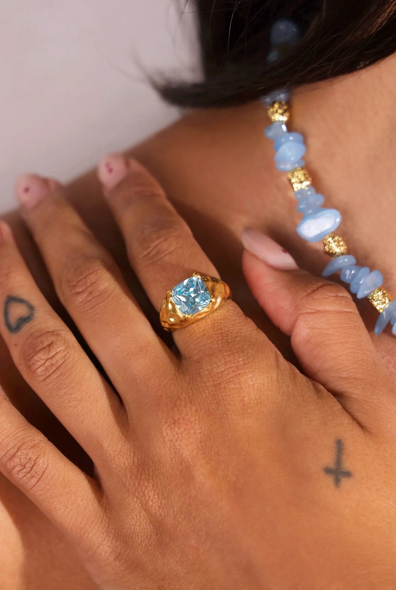 Close-up of a hand wearing a gold ring with a blue gemstone, with a beaded necklace in the background.