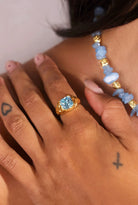 Close-up of a hand wearing a gold ring with a blue gemstone, with a beaded necklace in the background.