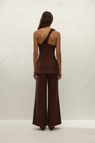 Person wearing a brown one-shoulder top and wide-leg pants against a white textured wall.