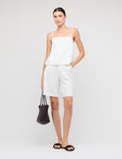 Woman wearing a white sleeveless top and shorts with a black handbag on a plain background