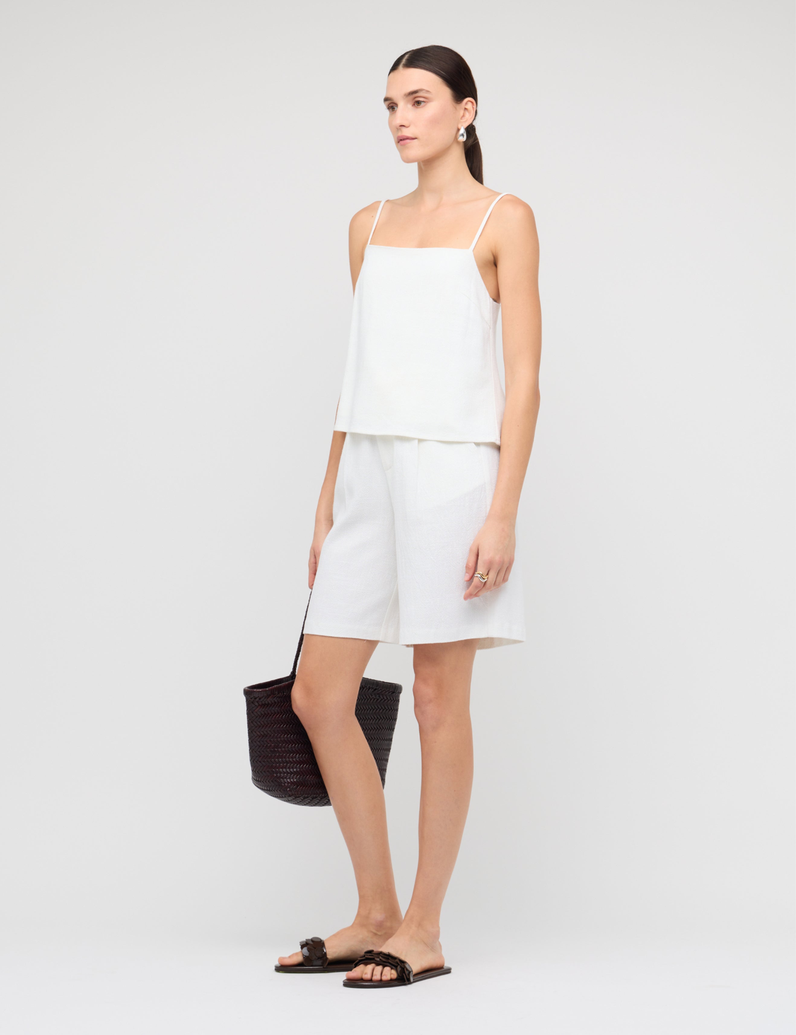 Woman wearing a white sleeveless top and shorts with a black handbag on a light gray background