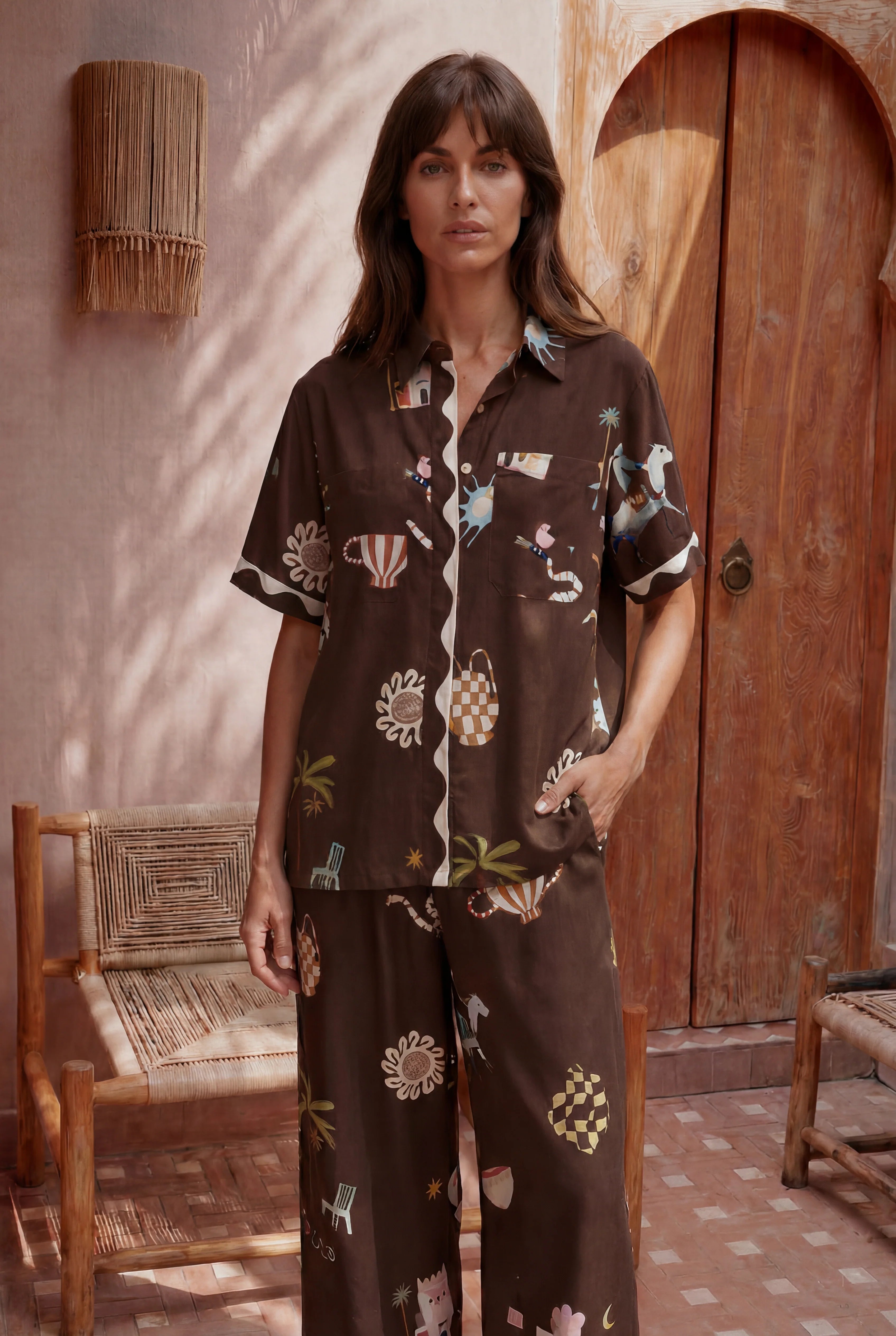 Woman wearing a patterned outfit standing in a sunlit room with wooden furniture.