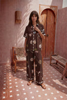 Woman wearing a floral outfit standing in a room with wooden furniture and a patterned floor.