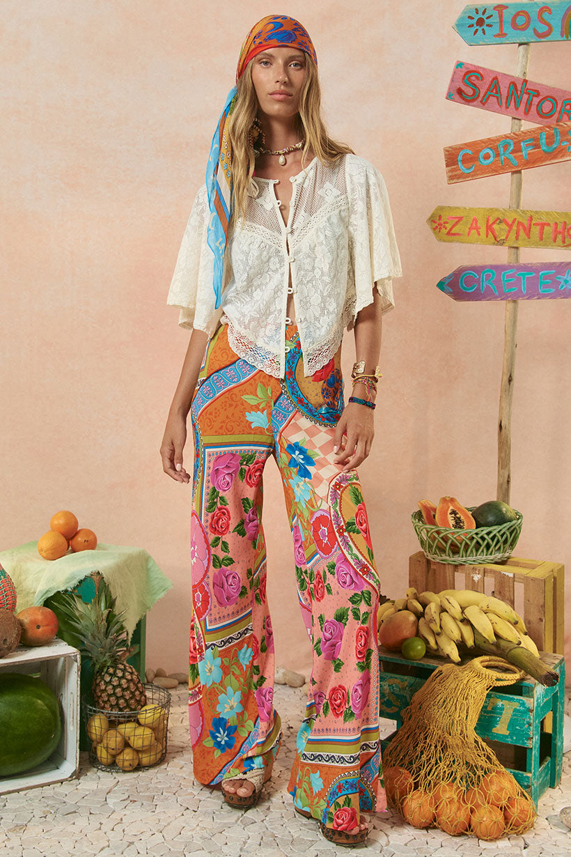 A model wearing a white shirt and pants with a colorful floral and geometric pattern. The setting includes decorative items and a signboard in the background.
