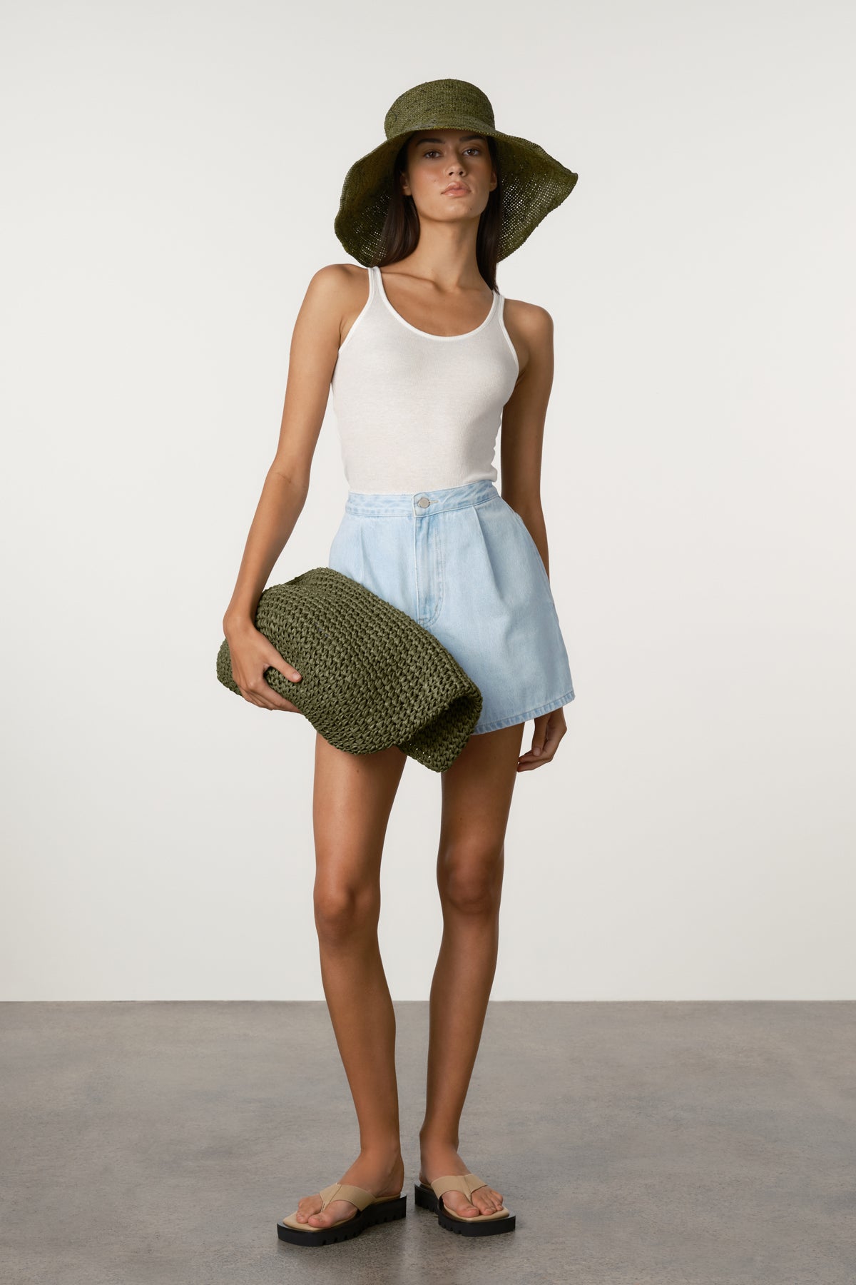Woman wearing a white tank top, light blue shorts, and a green hat holding a green clutch against a plain background.
