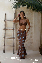 Woman wearing a brown bra and polka dot skirt standing in a sunlit room with a wooden ladder and plant.
