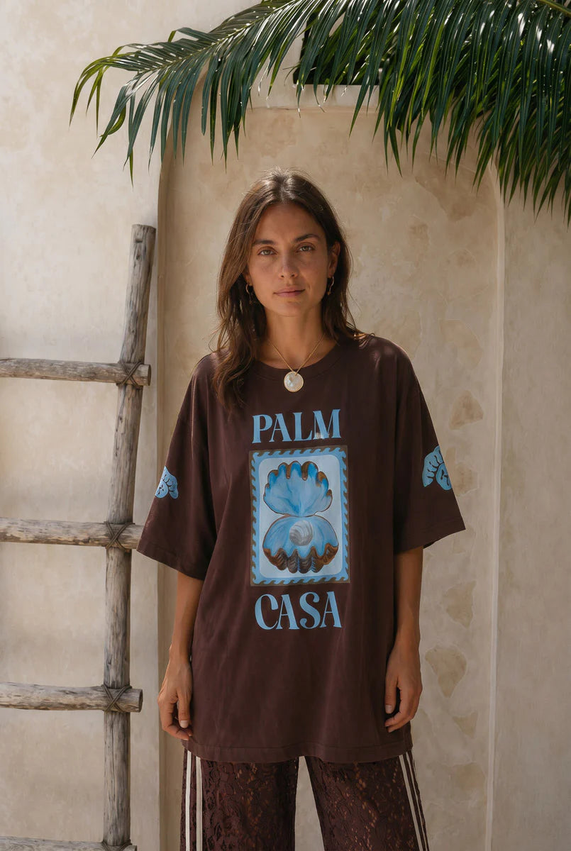Woman wearing a brown t-shirt with 'Palm Casa' design against a beige wall.