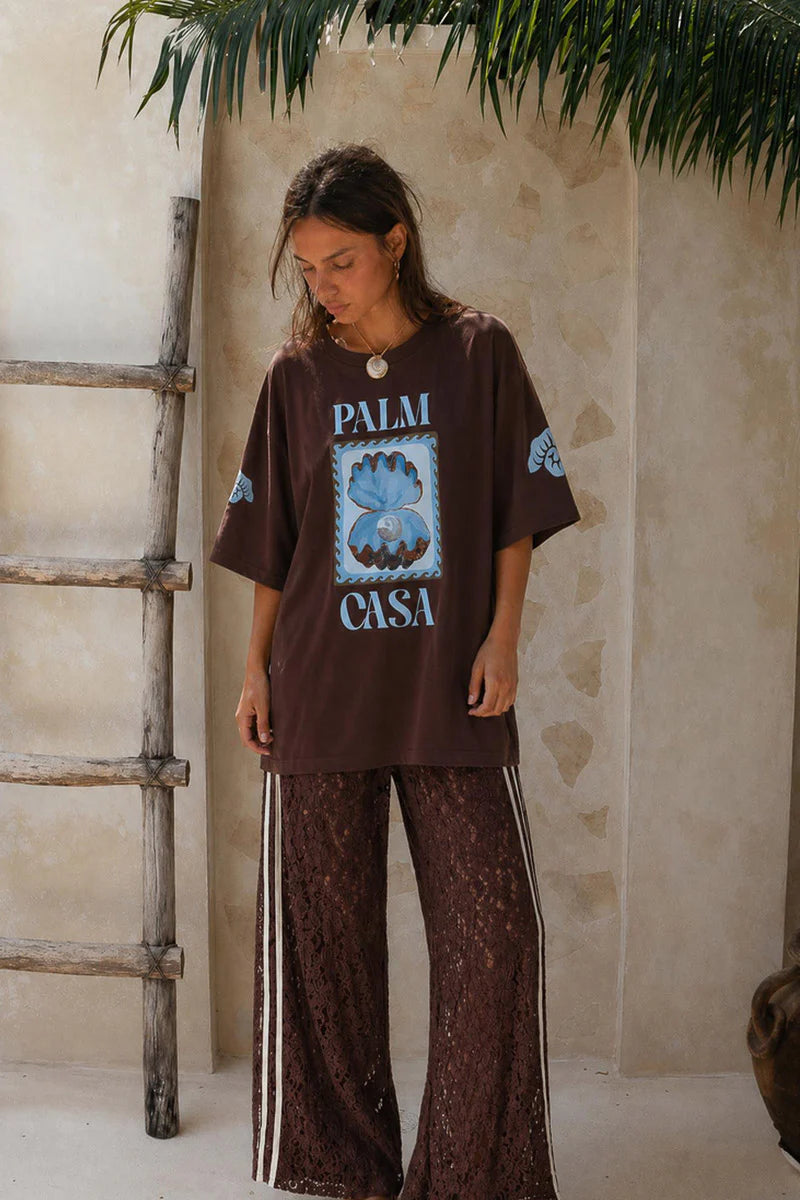 Person wearing a brown 'Palm Casa' t-shirt in a rustic setting with a ladder and plant.