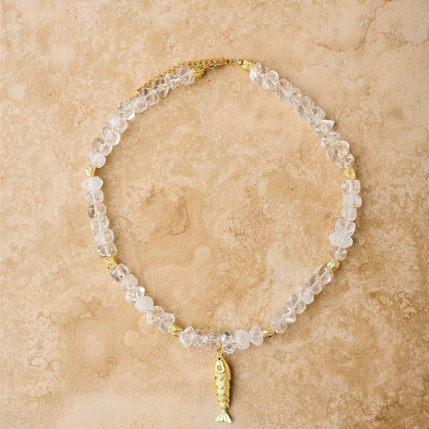 Necklace with clear beads and gold fish charm on a beige stone surface
