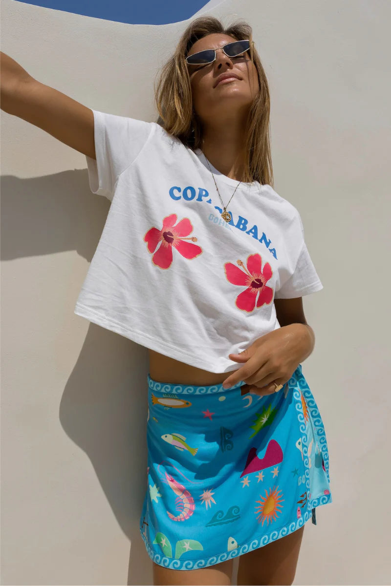 Woman wearing a white t-shirt with floral designs and 'Copa do Arara' text, paired with a blue skirt featuring colorful patterns.
