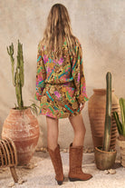 Person wearing a colorful patterned outfit with brown boots standing in front of potted plants and vases.