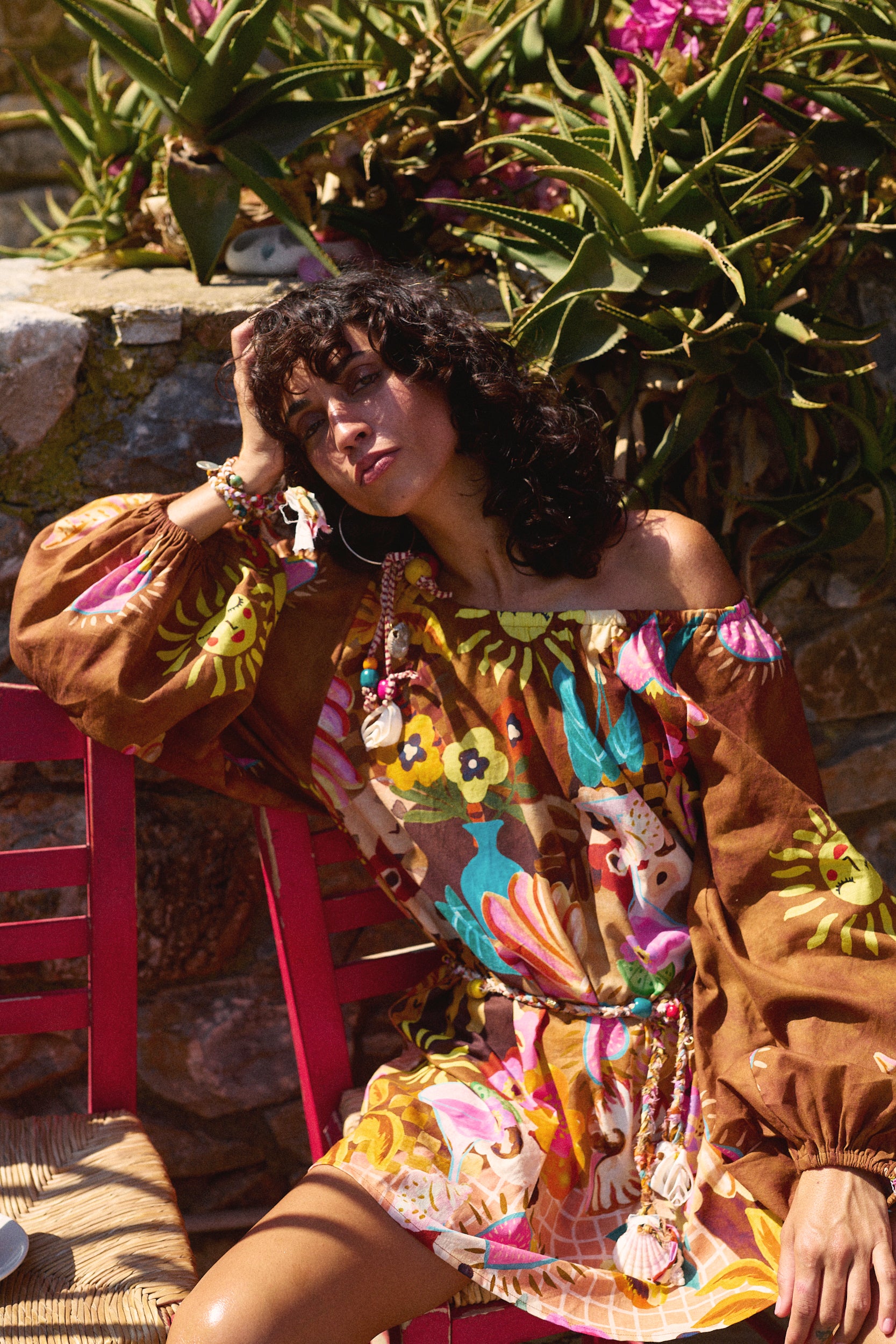 Woman in a colorful dress with floral patterns sitting outdoors near plants.