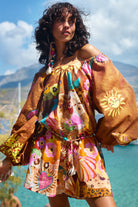 Woman wearing a colorful off-shoulder dress with floral patterns and a sun design, standing outdoors with mountains in the background.