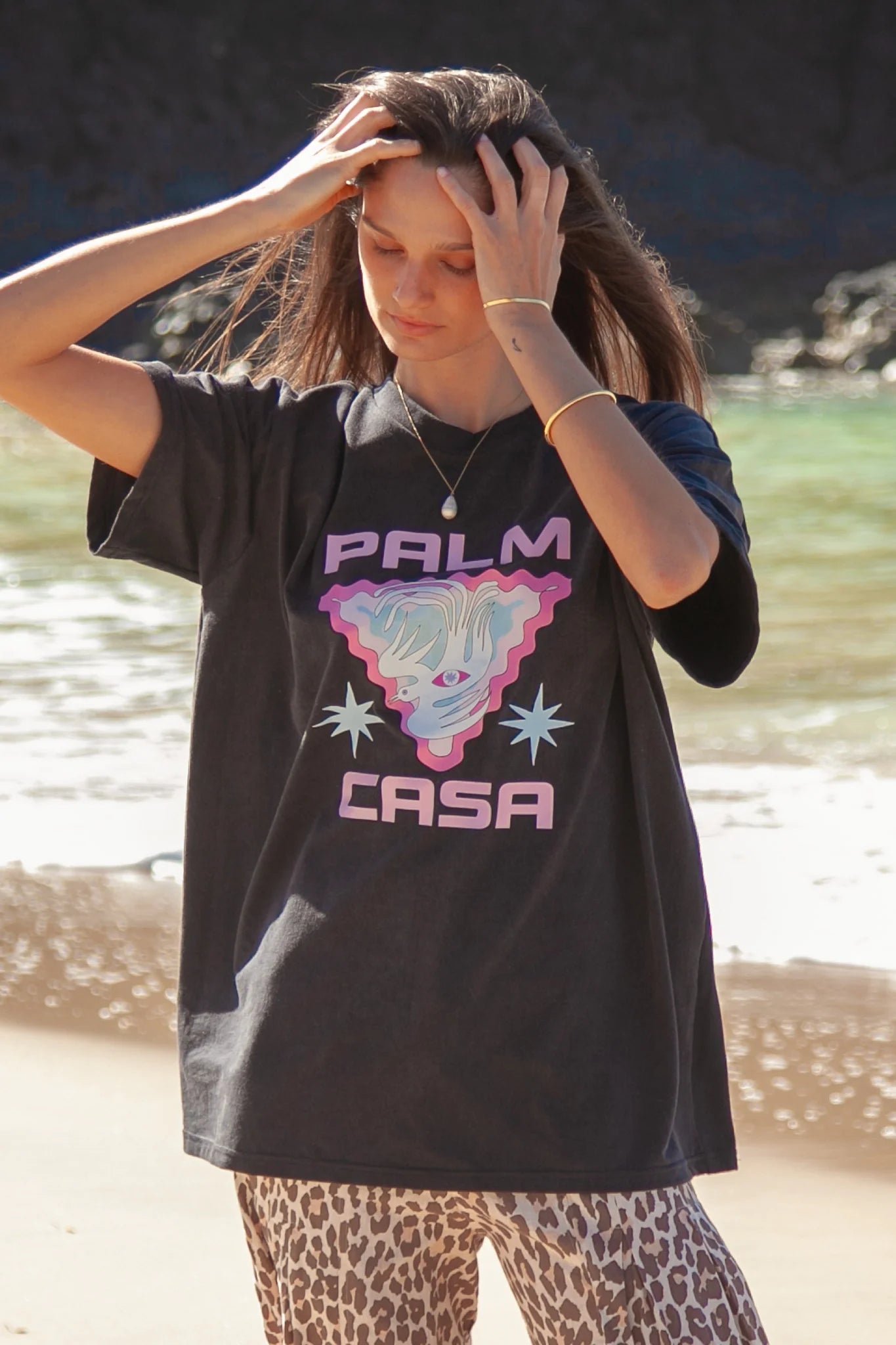 Person wearing a black t-shirt with 'Palm Casa' design by a body of water.