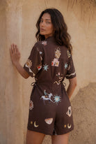 Woman wearing a brown romper with colorful patterns against a textured beige wall.