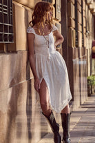 Woman in a white dress and black boots leaning against a wall in an urban setting.