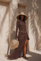 Woman in a patterned dress and wide-brimmed hat standing against a textured wall.