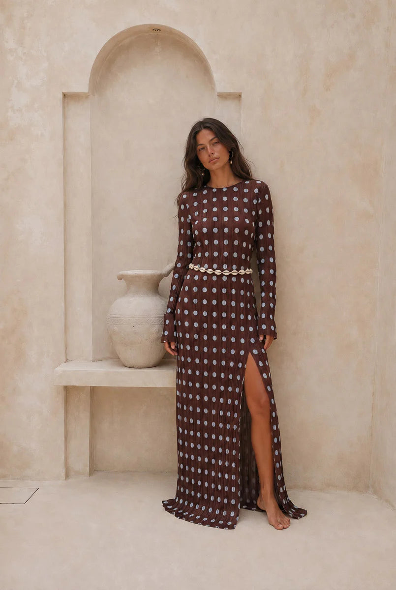 Woman in a patterned dress standing in a room with a stone wall and vase.