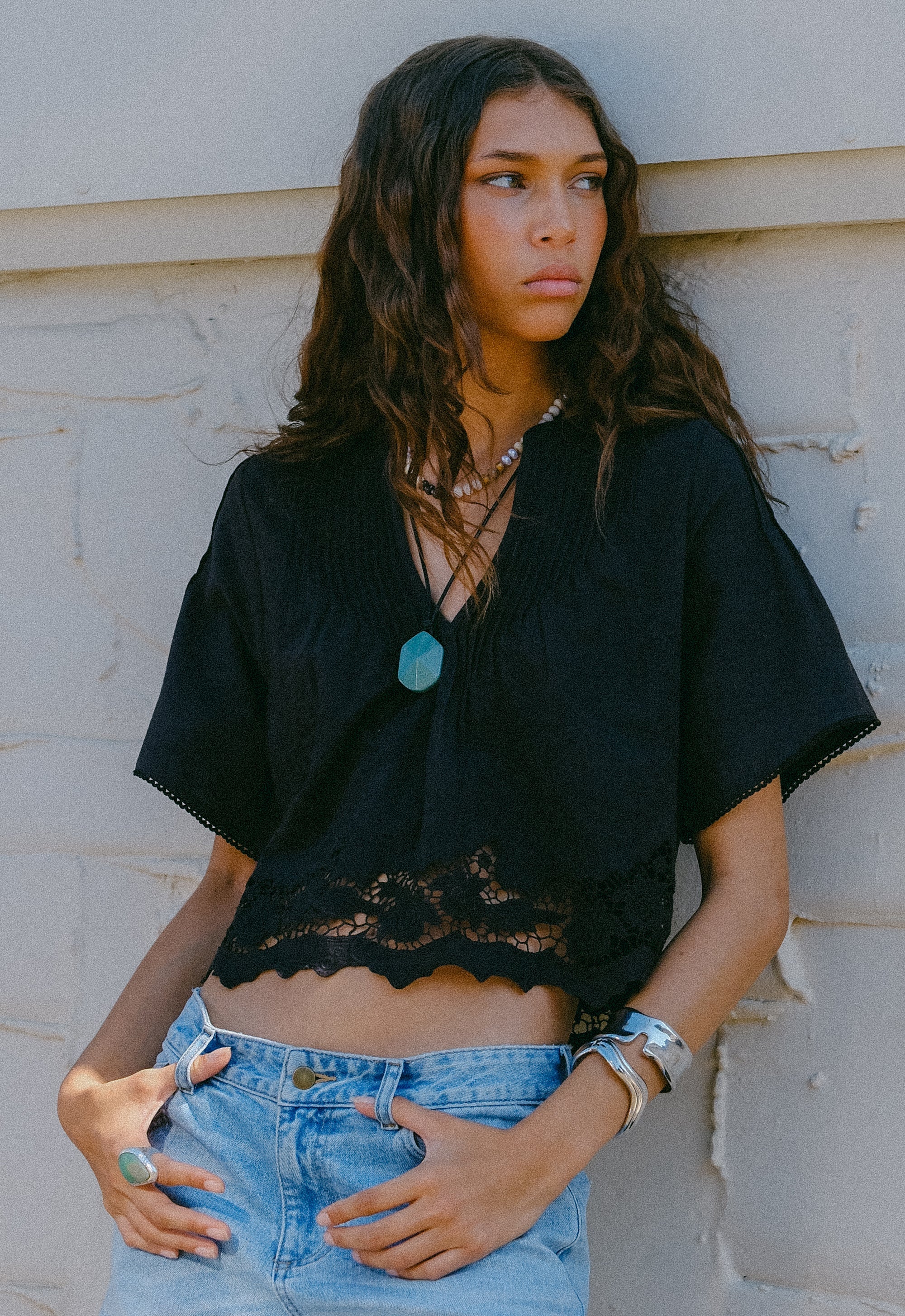 Woman wearing a black top with a lace detail and denim shorts against a light-colored wall.