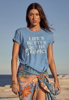 Woman wearing a blue t-shirt with 'Life's Better in the Tropics' text on a beach.