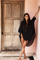 Woman in a black cover-up standing against a pink wall with a wooden door.
