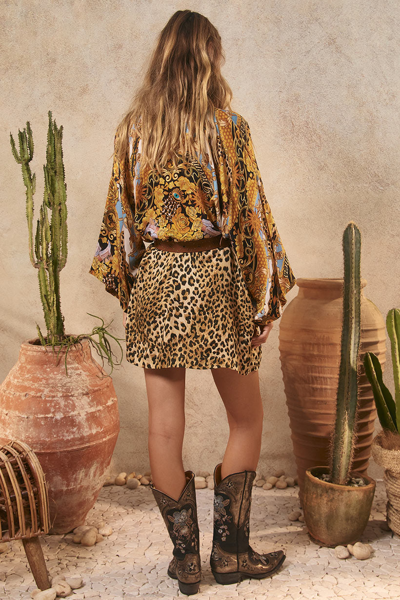 Person wearing a patterned shirt, leopard print skirt, and boots standing in a rustic setting with cacti and pots.