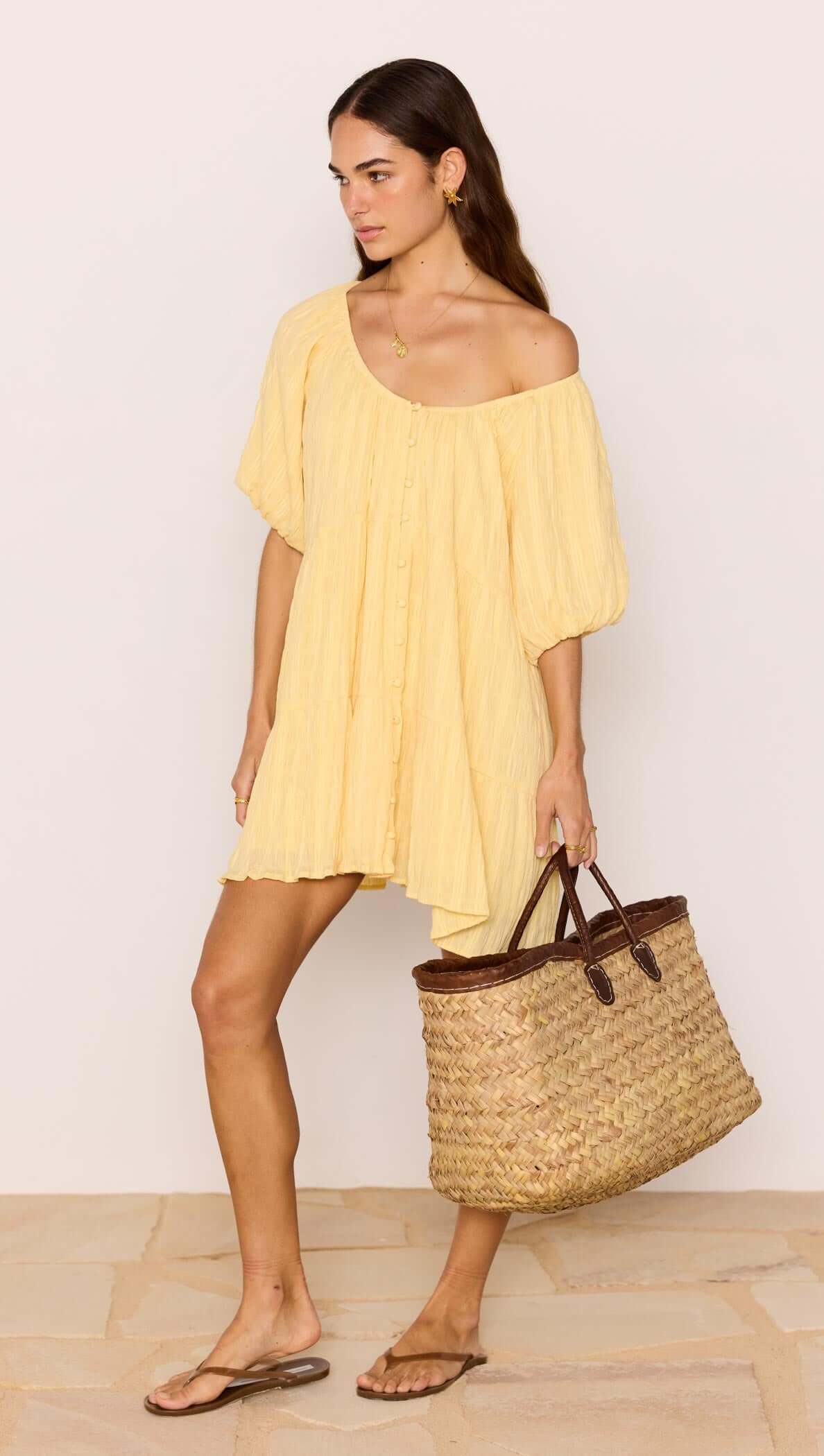 Woman in a yellow dress holding a straw bag against a plain background
