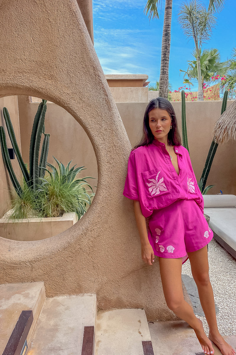 Woman in a pink outfit standing in front of a textured wall with plants and palm trees in the background.