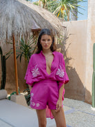 Woman in a pink outfit standing under a thatched umbrella with palm trees in the background