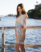 Woman in a colorful dress standing by a waterfront with trees in the background