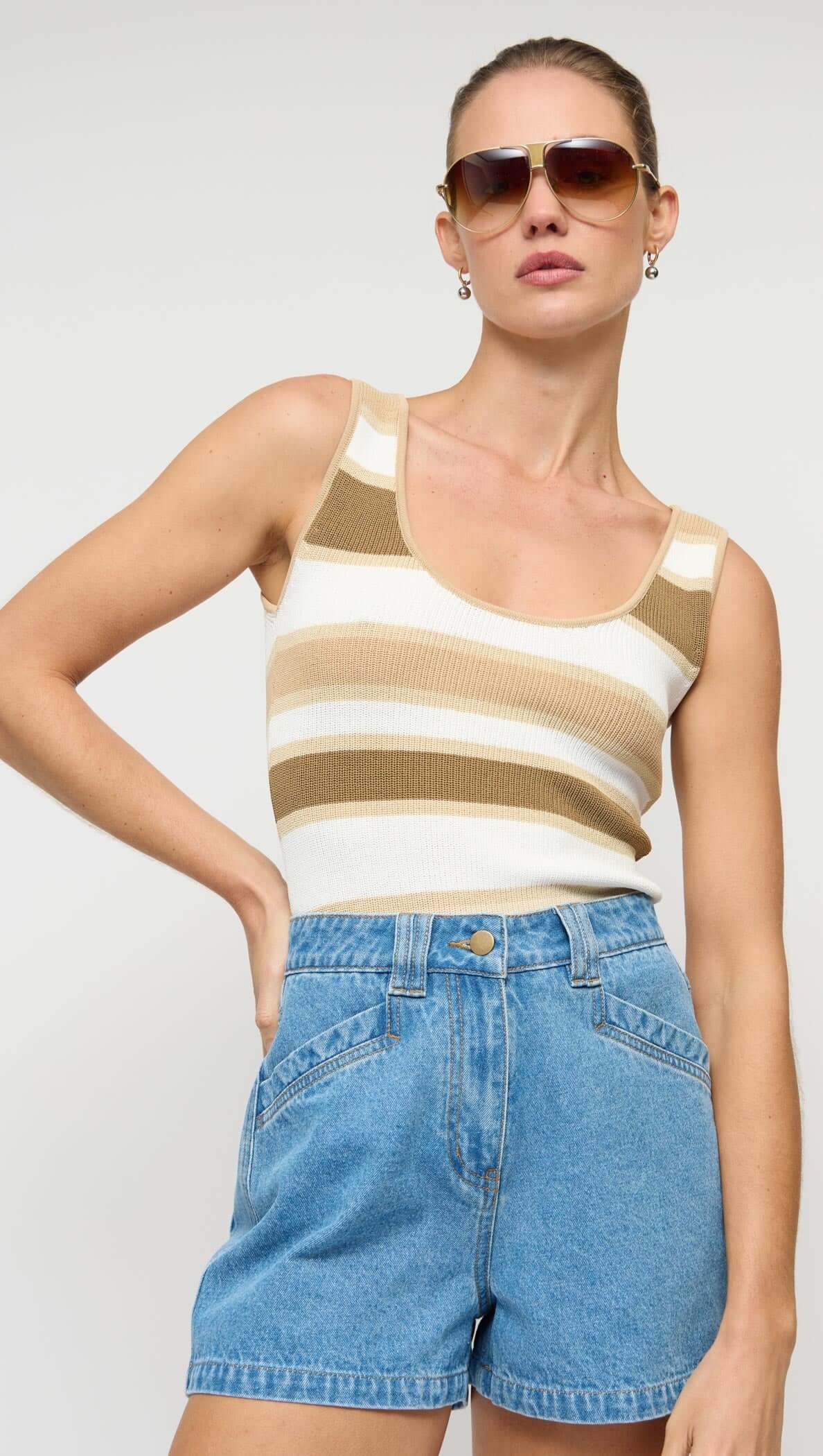 Woman wearing a striped tank top and denim shorts with sunglasses on a plain background