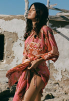 Woman in a pink and orange patterned dress standing in front of a rustic wall.