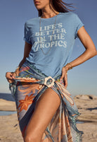Woman wearing a blue t-shirt with 'Life's Better in the Tropics' text on a beach.