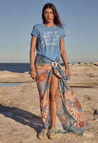 Woman on a beach wearing a blue t-shirt with 'Life's Better in the Tropics' text and a colorful sarong.