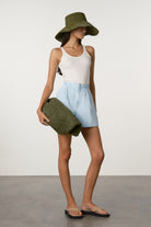 Woman wearing a white tank top, light blue shorts, and a green hat, holding a green clutch against a white background.