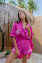 Woman in a pink outfit standing under a thatched umbrella with palm trees in the background
