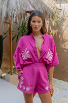 Woman wearing a bright pink outfit with floral patterns under a thatched umbrella.